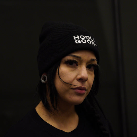 A woman wearing a black beanie with white 'HOOLIOON' text and a black t-shirt, photographed in a dark indoor setting.