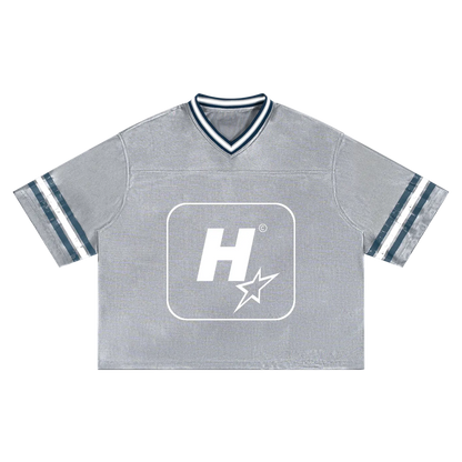 A heather gray cropped V-neck t-shirt with a white graphic logo on the chest and navy and white striped details on the collar and sleeves, shown flat on a white background.